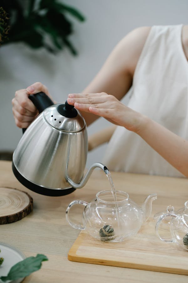 A woman is pouring hot water into a glass teapot from an electric kettle indoors.